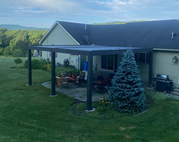 Black aluminum pergola with white louvered panels over patio seating with fire pit and brick home