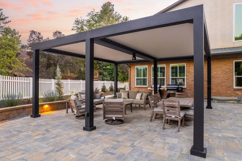 Black aluminum pergola with louvered roof over stone patio at sunset with outdoor furniture