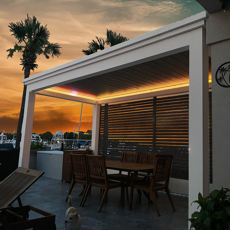Motorized pergola with warm LED lighting at dusk, white frame, dark louvered panels, furnished patio